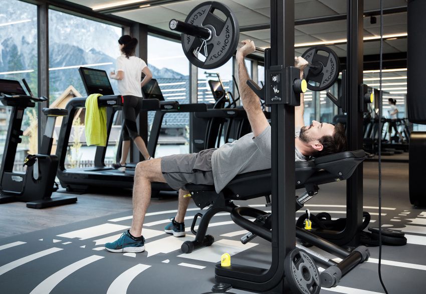 Gym with a woman in the background on a treadmill, in the foreground a man lifting weights.