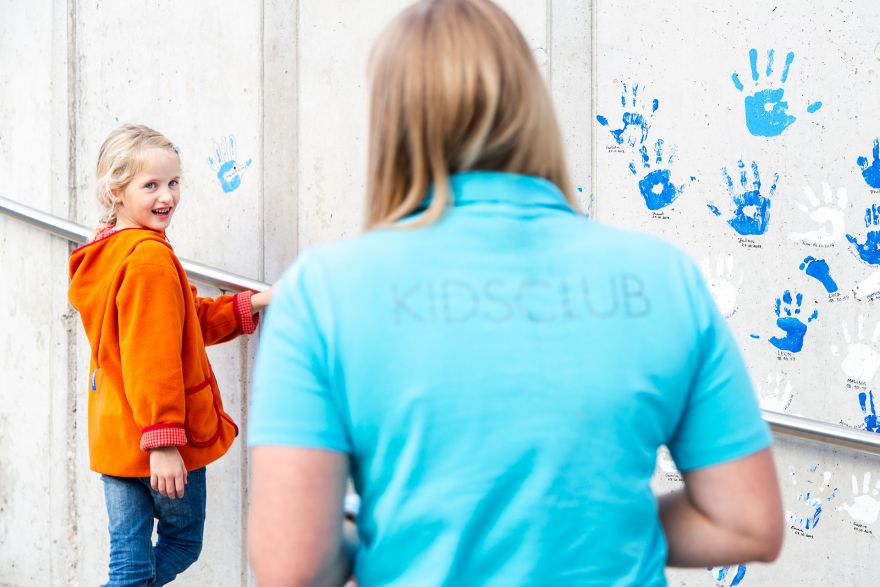 Ein junges Mädchen lächelt eine Frau im Vordergrund an, die ein hellblaues T-Shirt mit der Aufschrift 