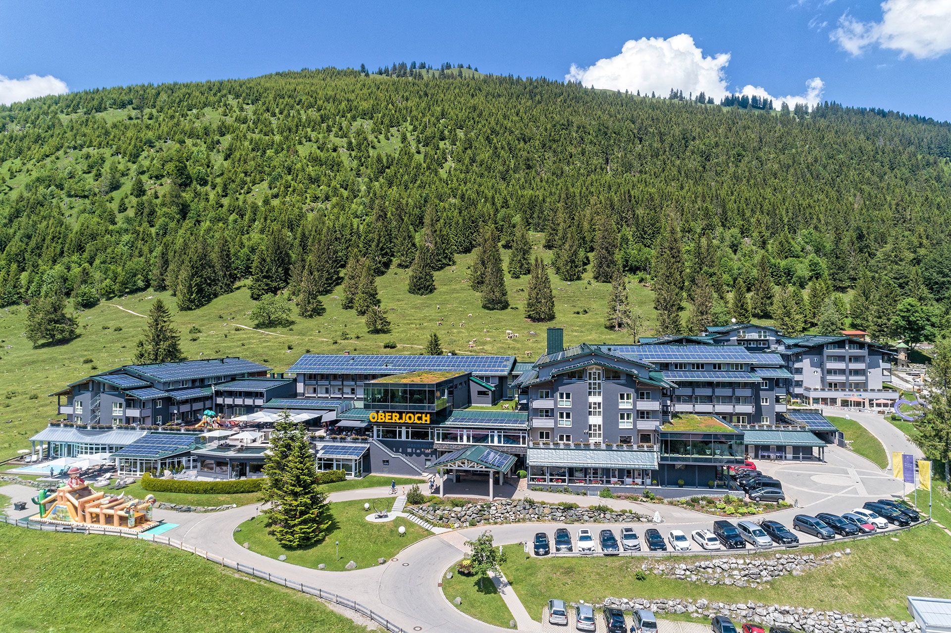 Das Kinderhotel Oberjoch aus der Vogelperspektive, im Hintergrund zahlreiche Bäume auf grüner Wiese.