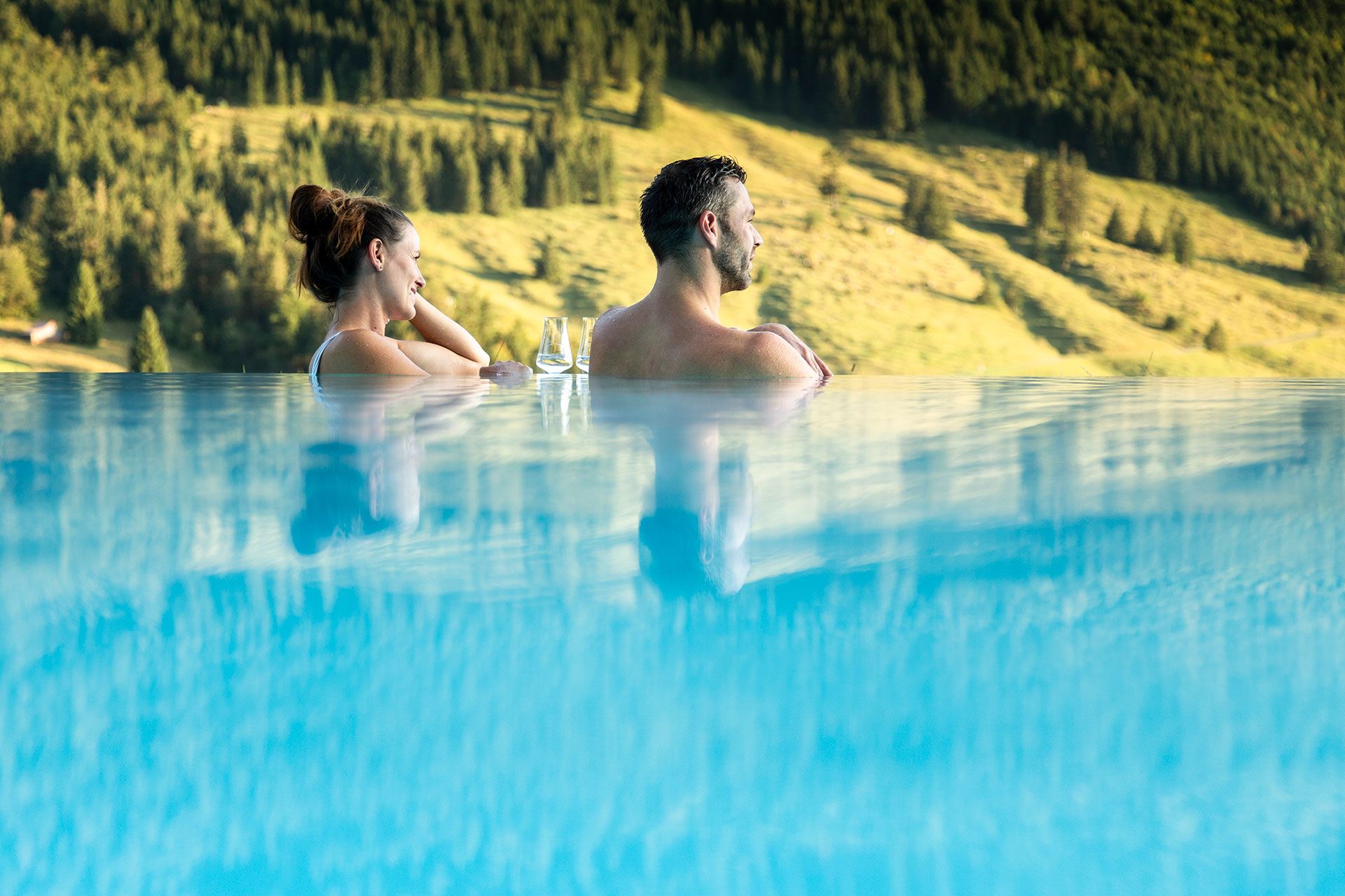Ein Mann und eine Frau blicken glücklich aus einem Outdoorpool, vor ihnen am Rand stehen zwei Weingläser. 