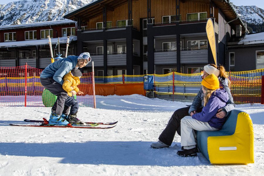 A man with his son between his legs in the snow, both on skis, to the right mother and daughter sitting on a colorful bench.